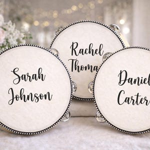 May include: Three tambourines with names on them. The tambourines have a cream-coloured surface with black trim and silver jingles. The names Sarah Johnson, Rachel Thomas, and Daniel Carter are written in black script.