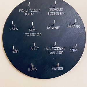 May include: A black circular game board with 10 hooks and corresponding text prompts for a drinking game. The text prompts include "Pick a Tosser to Sip", "Previous Tosser Sip", "2 Sips", "Next Tosser Sip", "Down It", "Skip a Go", "Shot", "1 Sip", "All Tossers Take a Sip", "3 Sips", "5 Sips", and "Water".