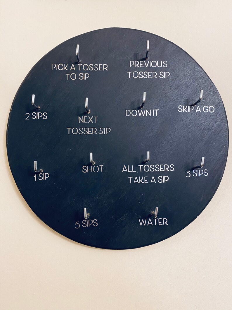 May include: A black circular game board with 10 hooks and corresponding text prompts for a drinking game. The text prompts include "Pick a Tosser to Sip", "Previous Tosser Sip", "2 Sips", "Next Tosser Sip", "Down It", "Skip a Go", "Shot", "1 Sip", "All Tossers Take a Sip", "3 Sips", "5 Sips", and "Water".