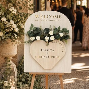 May include: An arched wooden wedding welcome sign on a tripod easel. The sign features the text "WELCOME TO THE WEDDING OF" and a heart-shaped cutout displaying "JESSICA & CHRISTOPHER" with the date "03/08/2024" and artificial white roses and greenery.