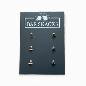 May include: A dark gray chalkboard sign with the text "BAR SNACKS" and a graphic of two beer mugs. Six silver clips are attached to the board.