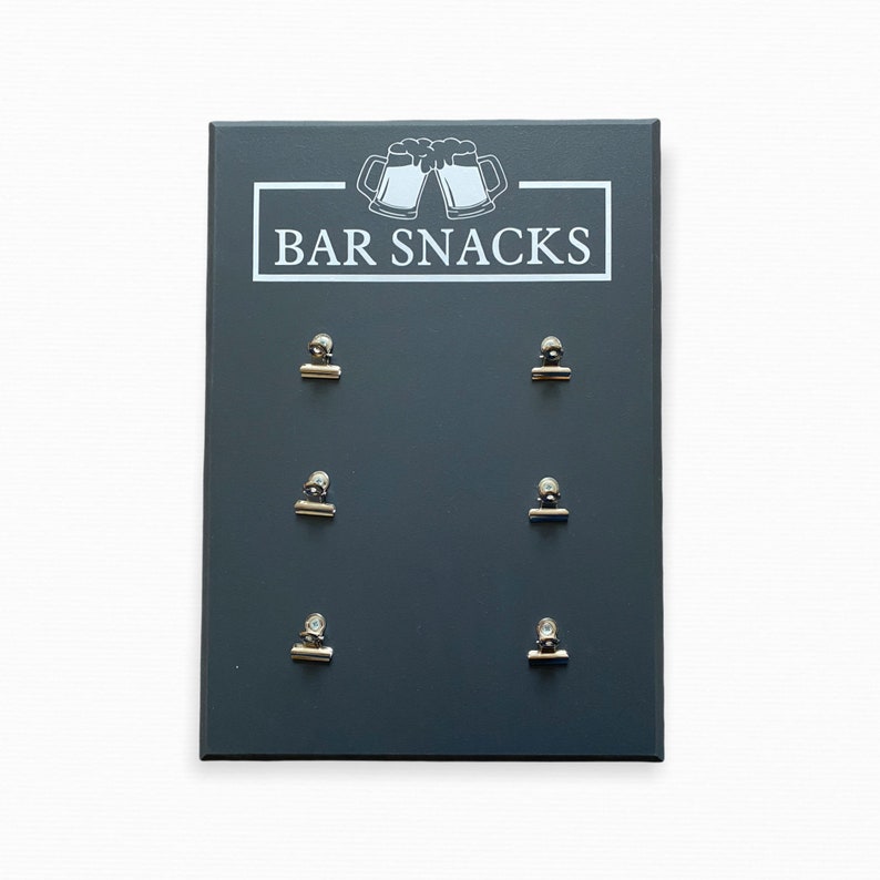 May include: A dark gray chalkboard sign with the text "BAR SNACKS" and a graphic of two beer mugs. Six silver clips are attached to the board.