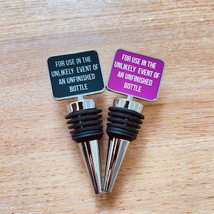 May include: Two bottle stoppers with the text "FOR USE IN THE UNLIKELY EVENT OF AN UNFINISHED BOTTLE". One stopper has a black background, the other is magenta. Both have a silver base and black rubber seal.