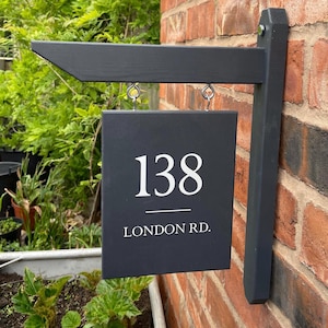 May include: A dark grey rectangular hanging sign mounted on a brick wall. The sign features the white text 138 above the text LONDON RD. with a horizontal line separating them. The sign is suspended from a dark grey wooden arm by two metal hooks.