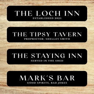 May include: Four black rectangular signs with white text on a wood grain background. The signs read: "THE LOCH INN ESTABLISHED 2025", "THE TIPSY TAVERN PROPRIETOR: SHELLEY SMITH", "THE STAYING INN SERVED IN THE SHED", and "MARK'S BAR GOOD SPIRITS, BAD JOKES".
