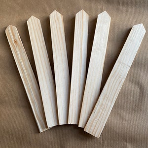 May include: Seven wooden plant markers with pointed tops, arranged in a fan shape on a brown paper background.