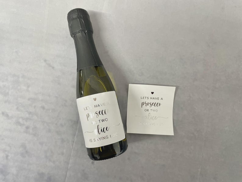 May include: A small glass bottle with a black foil neck wrap and a white label featuring the text "LETS HAVE A PROSECCO OR TWO ALICE IS SAYING I DO" with a small heart icon. Beside the bottle is a matching square white sticker with the same text.