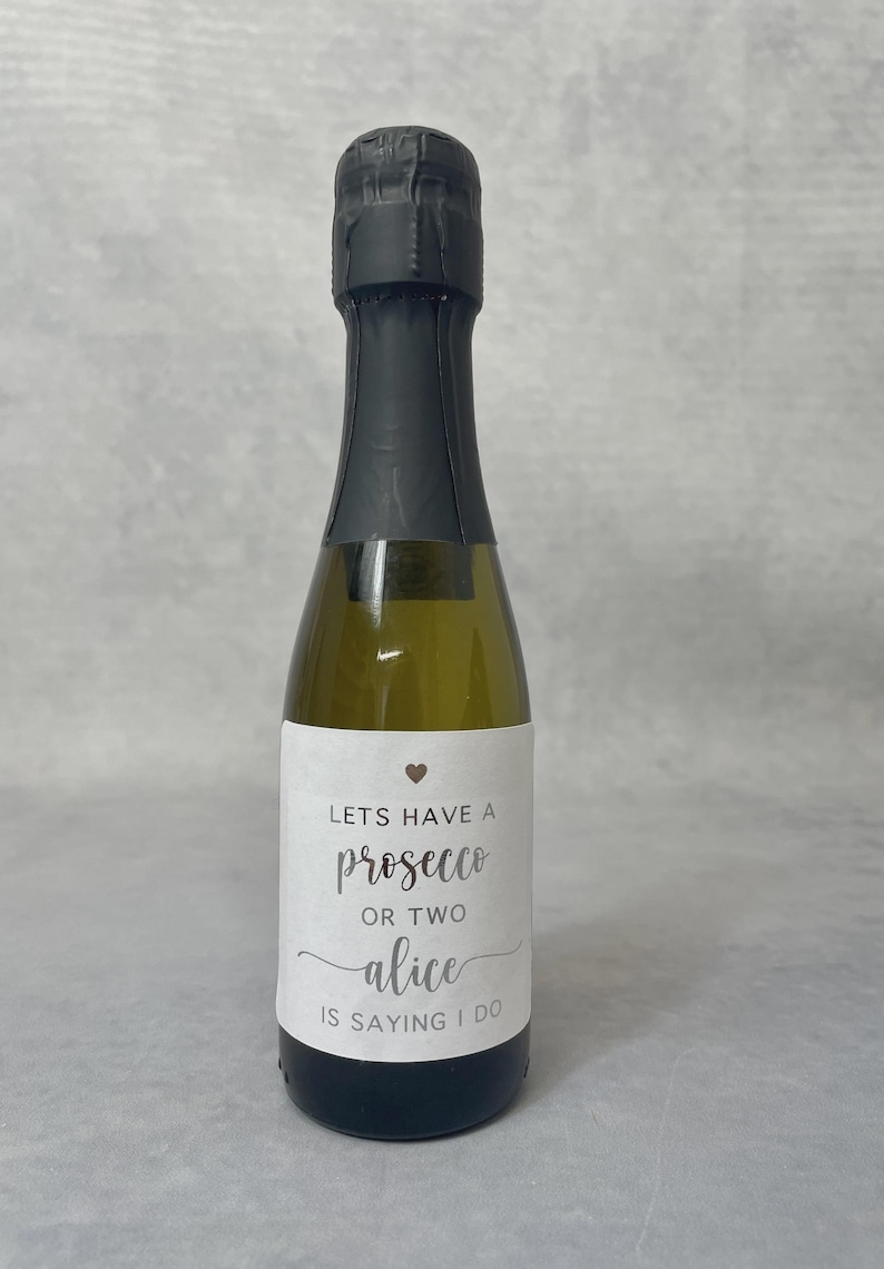 May include: A small glass bottle of prosecco with a black foil-wrapped neck and a white rectangular label. The label features a small heart icon and the text: LETS HAVE A prosecco OR TWO alice IS SAYING I DO in black and grey lettering.
