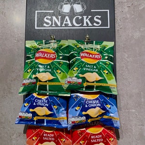 May include: A black chalkboard sign with the word "SNACKS" written in white. Six bags of Walker's crisps are displayed on the chalkboard. The crisps are in three flavors: Salt & Vinegar, Cheese & Onion, and Ready Salted.