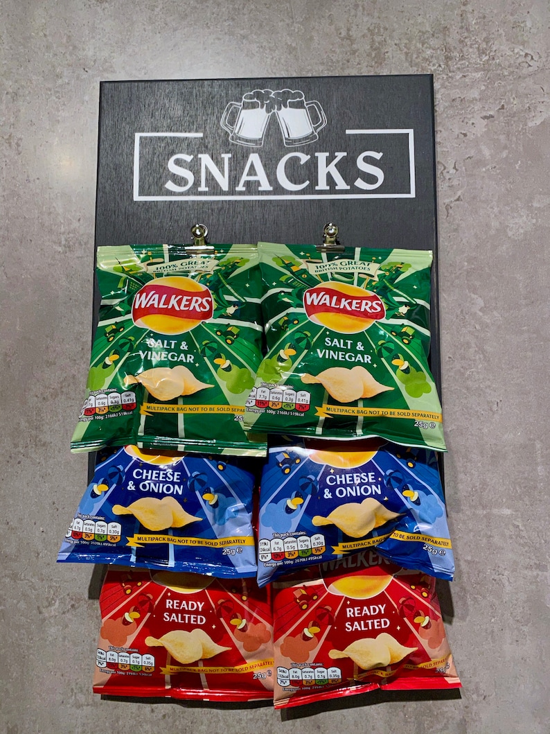 May include: A black chalkboard sign with the word "SNACKS" written in white. Six bags of Walker's crisps are displayed on the chalkboard. The crisps are in three flavors: Salt & Vinegar, Cheese & Onion, and Ready Salted.