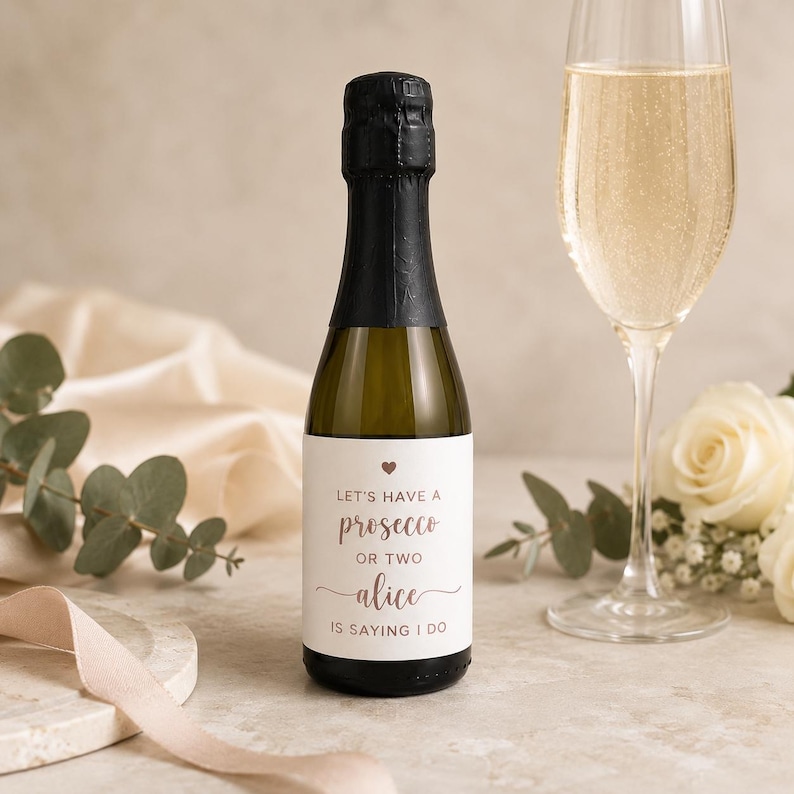 May include: A small bottle of prosecco with a white label featuring a small heart icon and the text: LET'S HAVE A prosecco OR TWO alice IS SAYING I DO. The bottle has a black foil-wrapped neck and is positioned next to a tall, clear glass flute.