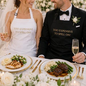 Puede incluir: Una novia y un novio en una recepción de boda, ambos con delantales con el texto "TOO EXPENSIVE TO SPILL ON!". El delantal de la novia es blanco, el del novio es negro. Están sentados a la mesa con comida y champán.