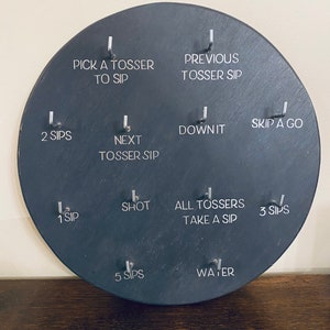 May include: A black circular slate board with 10 different drinking game rules written in white text. The rules include "Pick a Tosser to Sip", "Previous Tosser Sip", "2 Sips", "Next Tosser Sip", "Down It", "1 Sip", "Shot", "All Tossers Take a Sip", "5 Sips", "Water", "Skip a Go", and "3 Sips".