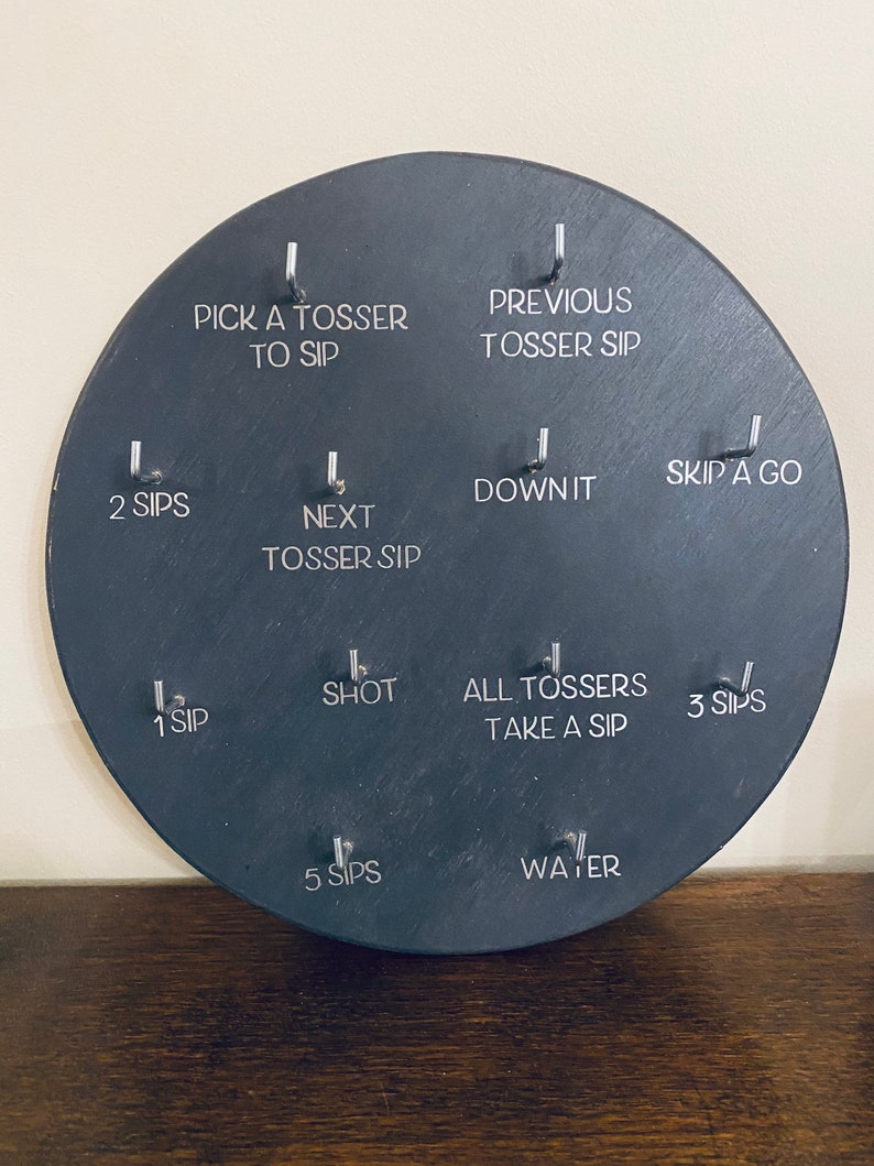 May include: A black circular slate board with 10 different drinking game rules written in white text. The rules include "Pick a Tosser to Sip", "Previous Tosser Sip", "2 Sips", "Next Tosser Sip", "Down It", "1 Sip", "Shot", "All Tossers Take a Sip", "5 Sips", "Water", "Skip a Go", and "3 Sips".