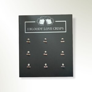 May include: A black sign with the text "I Bloody Love Crisps" and a graphic of two beer mugs clinking. Nine silver binder clips are attached to the sign.