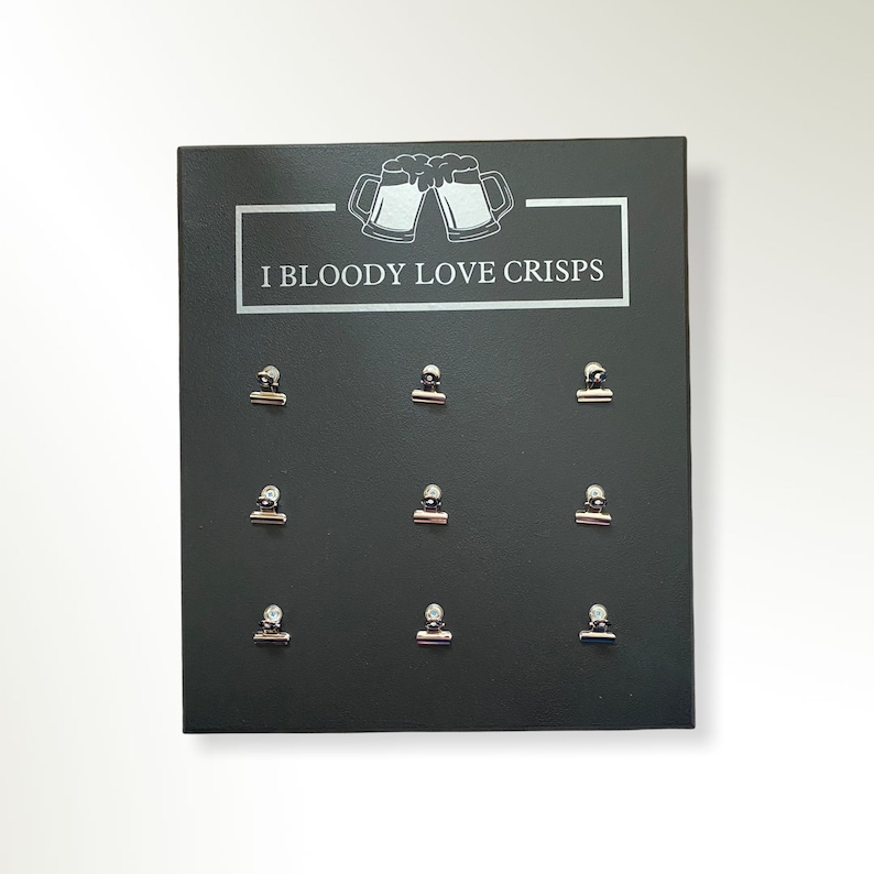 May include: A black sign with the text "I Bloody Love Crisps" and a graphic of two beer mugs clinking. Nine silver binder clips are attached to the sign.