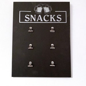 May include: Black chalkboard sign with the word "SNACKS" in white lettering and a graphic of two beer mugs. Six silver binder clips are attached to the board.