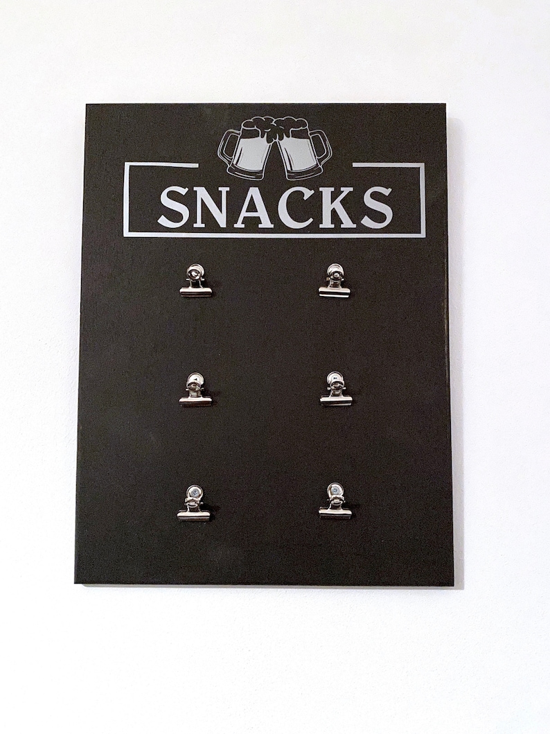 May include: Black chalkboard sign with the word "SNACKS" in white lettering and a graphic of two beer mugs. Six silver binder clips are attached to the board.