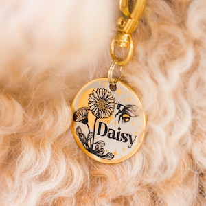 May include: Gold dog tag with a black and white illustration of a daisy and a bee. The tag says "Daisy" in black lettering.