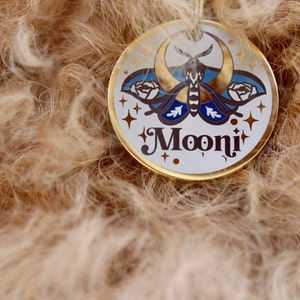 Puede incluir: Una chapa redonda para perro en blanco y dorado con una luna creciente, una polilla y dos rosas. La chapa dice "Mooni" en una fuente estilizada.
