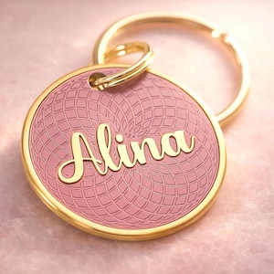 以下が含まれることがあります： 筆記体で「Alina」と書かれた名前入りのゴールドカラーのキーホルダー。 ピンク色のペンダントにはテクスチャパターンとゴールドの縁取りがあり、ゴールドのリングに付いています。