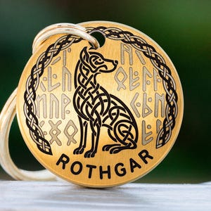 May include: A circular, gold-toned dog tag featuring a black Celtic dog design and the name "ROTHGAR". The tag has a braided border and runes. A cream-coloured cord is attached to the top. The tag is likely made of metal.