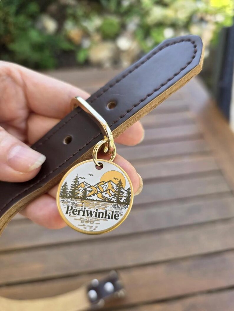 May include: A brown leather dog collar with a gold-colored metal ring and a circular tag. The tag features a mountain and tree design with the word "Periwinkle" in a serif font. The collar is held by a hand.