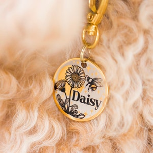May include: Gold dog tag with a black and white illustration of a daisy and a bee. The tag says "Daisy" in black lettering.