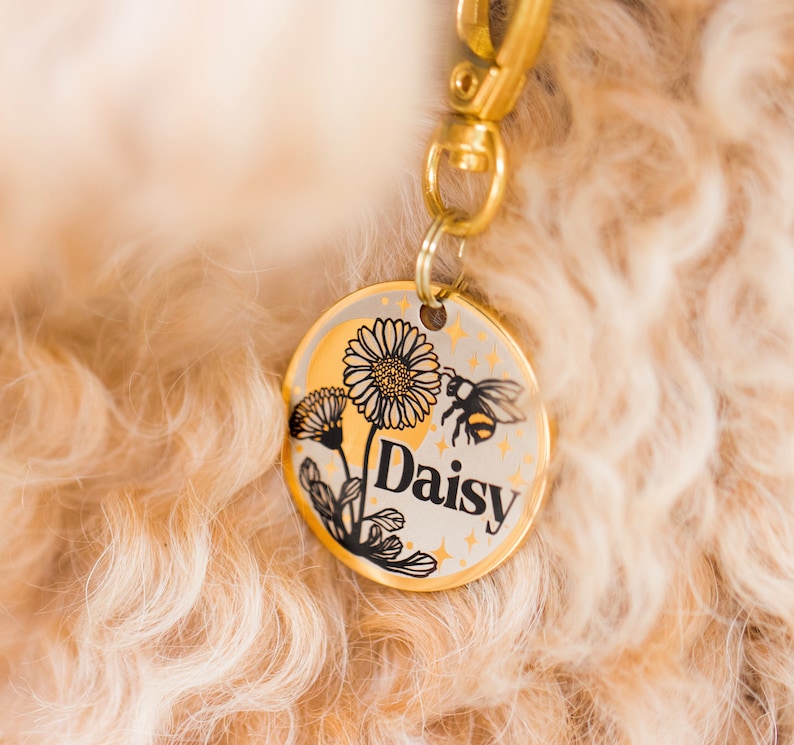 May include: Gold dog tag with a black and white illustration of a daisy and a bee. The tag says "Daisy" in black lettering.