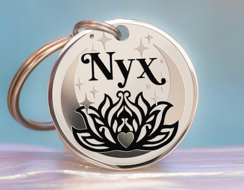 Puede incluir: Chapa de identificaci&oacute;n para mascotas de plata con una luna creciente negra, estrellas y un dise&ntilde;o de flor de loto estilizado. El nombre "Nyx" est&aacute; grabado en letras negras.