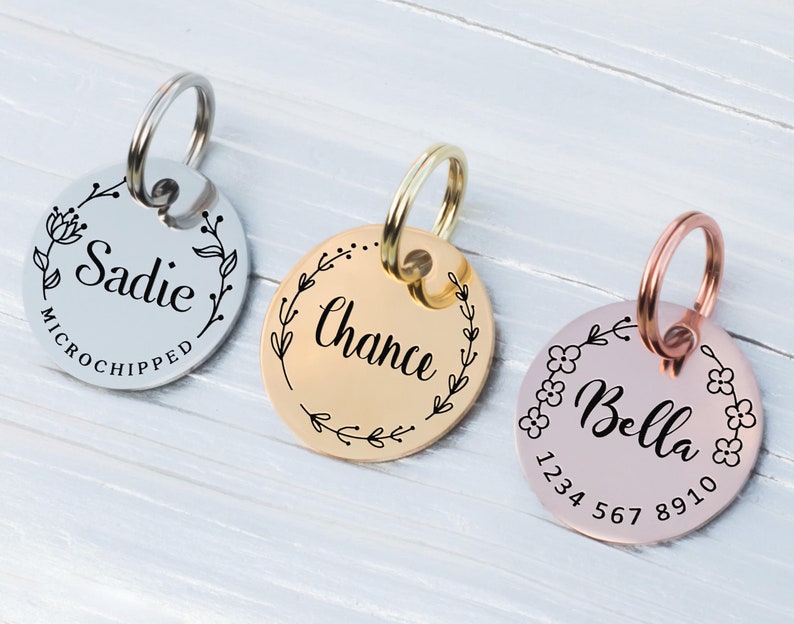 May include: Three personalized pet ID tags with floral designs. The tags are silver, gold, and rose gold, each with a different name: "Sadie", "Chance", and "Bella". The "Bella" tag also includes a phone number. All tags have a ring for attachment.