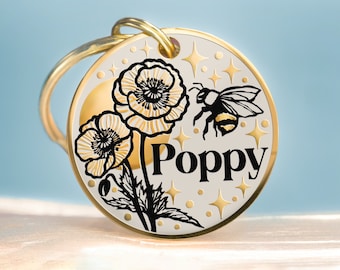 May include: Gold-toned metal pet tag with a white background featuring a black and white illustration of two poppies and a bee. The tag has the name "Poppy" written in black lettering.