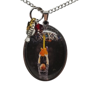May include: A silver oval pendant necklace with a photo of a person holding a guitar in front of a crowd. The pendant has a small silver bird charm with a red crystal.