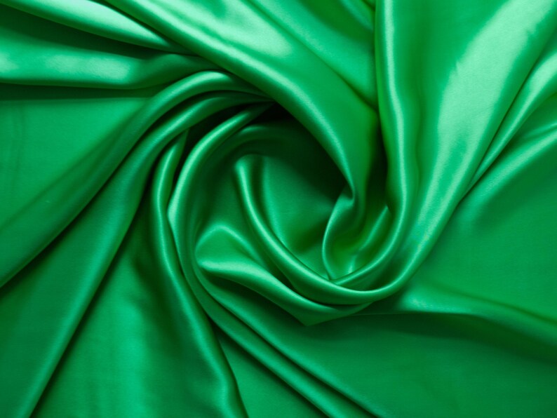 May include: A close-up shot of emerald green satin fabric. The fabric is draped and swirled, creating soft folds and shadows. The shiny material reflects light, highlighting the smooth texture. This textile is suitable for apparel and home decor.