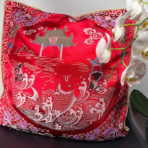 Könnte beinhalten: Rotes Zierkissen mit einem gestickten asiatisch inspirierten Design. Das Kissen zeigt ein zentrales Motiv eines Fisches im Wasser, mit Pagoden und Wolken. Die Ränder sind mit goldenem Kordel und floralen Mustern verziert.