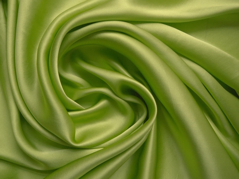 May include: Close-up of a vibrant, lime green fabric with a silky sheen. The material is draped and swirled, creating soft folds and shadows. The fabric's smooth texture and bright color suggest it could be used for apparel or home decor.