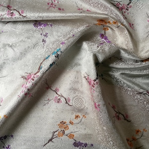 May include: A shimmering fabric with a floral pattern. The ivory-colored material features embroidered branches with pink, orange, purple, and blue blossoms. The fabric has a subtle embossed design and is draped in soft folds.