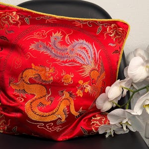 Könnte beinhalten: Rotes Zierkissen mit einem goldenen Drachen- und Phönix-Design. Das Kissen hat eine goldene Kordelbordüre und liegt auf einem schwarzen Stuhl. Weiße Orchideen sind im Hintergrund.
