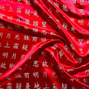 K&ouml;nnte beinhalten: Ein leuchtend roter Satin-Stoff mit flie&szlig;ender, drapierter Textur. Der Stoff ist mit silbernen chinesischen Schriftzeichen verziert, die ein Muster auf der Oberfl&auml;che bilden. Die Beleuchtung hebt die Falten und den Glanz des Materials hervor.