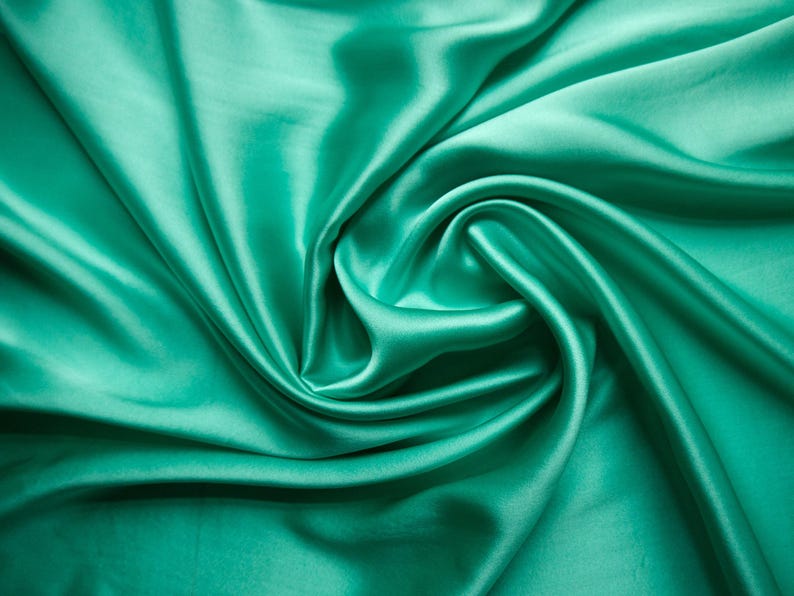 May include: Close-up of a vibrant, flowing fabric in a teal green hue. The material is smooth and lustrous, with soft folds and a swirling pattern. The fabric's sheen suggests a satin or silk texture, ideal for apparel or home decor.