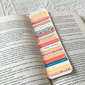 Könnte beinhalten: Ein farbenfroher Lesezeichen mit Buchrücken-Designs, darunter Titel wie "Le Dormeur du Val" und "Liberté". Es liegt auf einem Buch mit französischem Text, ideal für Leser und Bücherliebhaber.
