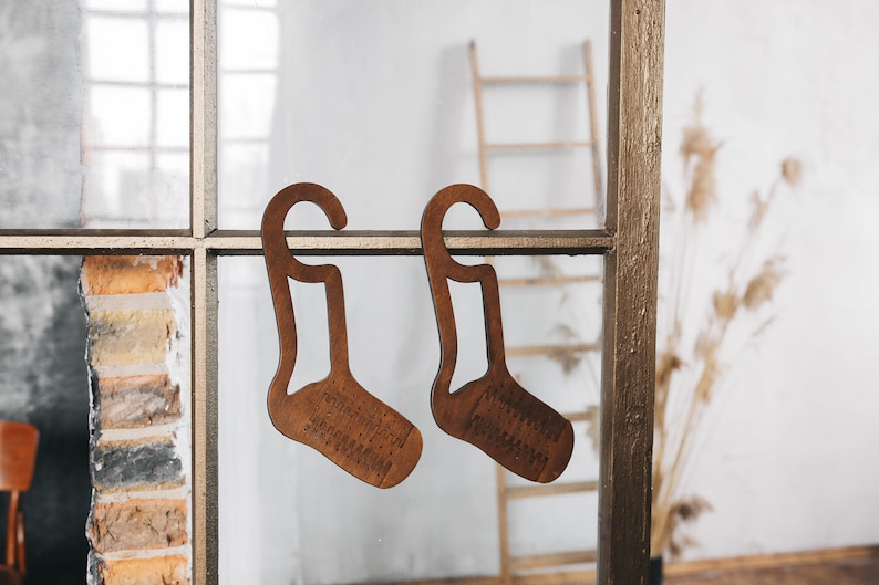 May include: Two wooden sock holders shaped like socks hanging on a window frame. The holders are brown and have a hook at the top.