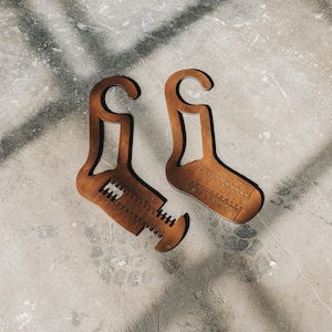 May include: Two brown wooden sock shapers with a hook at the top and a measurement scale on the bottom. The shapers are on a light grey concrete surface.