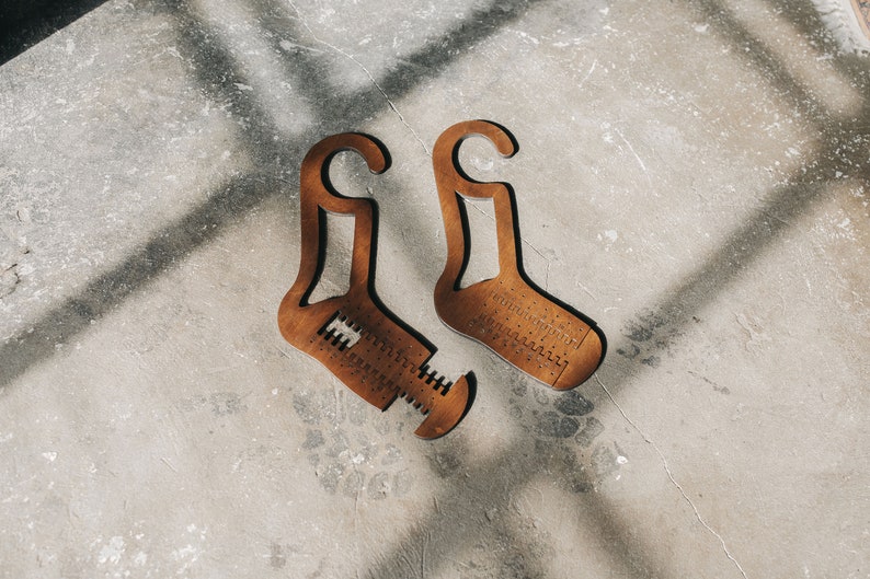 May include: Two brown wooden sock shapers with a hook at the top and a measurement scale on the bottom. The shapers are on a light grey concrete surface.