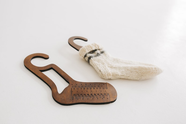 May include: Two wooden sock knitting tools with a hook at the top and a numbered scale on the bottom. One tool has a white and black striped sock hanging on it.