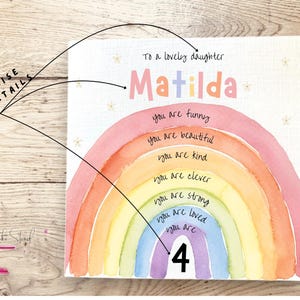 May include: A square white sign featuring a watercolor rainbow with the text To a lovely daughter, Matilda, and the phrases you are funny, you are beautiful, you are kind, you are clever, you are strong, you are loved, you are 4.