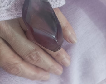 May include: A large, faceted, purple gemstone ring. The ring's stone is an elongated, rectangular shape with multiple facets, creating a sparkling effect. The ring is set on a hand, with a soft, light purple fabric in the background.