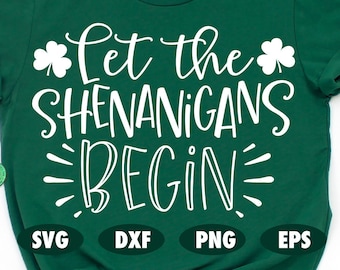 May include: Green t-shirt with white text that reads "Let the Shenanigans Begin". The shirt has shamrock designs on either side of the text. The shirt is laying flat on a white surface with green shamrock designs scattered around it.