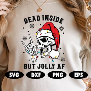 May include: A white sweatshirt with the text "Dead Inside But Jolly AF" in black lettering. A skeleton wearing a Santa hat and holding up a rock and roll hand gesture is in the center of the text. The skeleton is surrounded by Christmas lights and snowflakes. The words "SVG", "DXF", "PNG", and "EPS" are in black circles at the bottom of the sweatshirt.