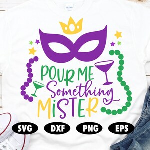 Op de afbeelding: Wit T-shirt met een paars masker, groene kralen en de tekst "POUR ME SOMETHING MISTER" in een groen en geel lettertype. Het shirt heeft ook een kroon en een martini-glas.